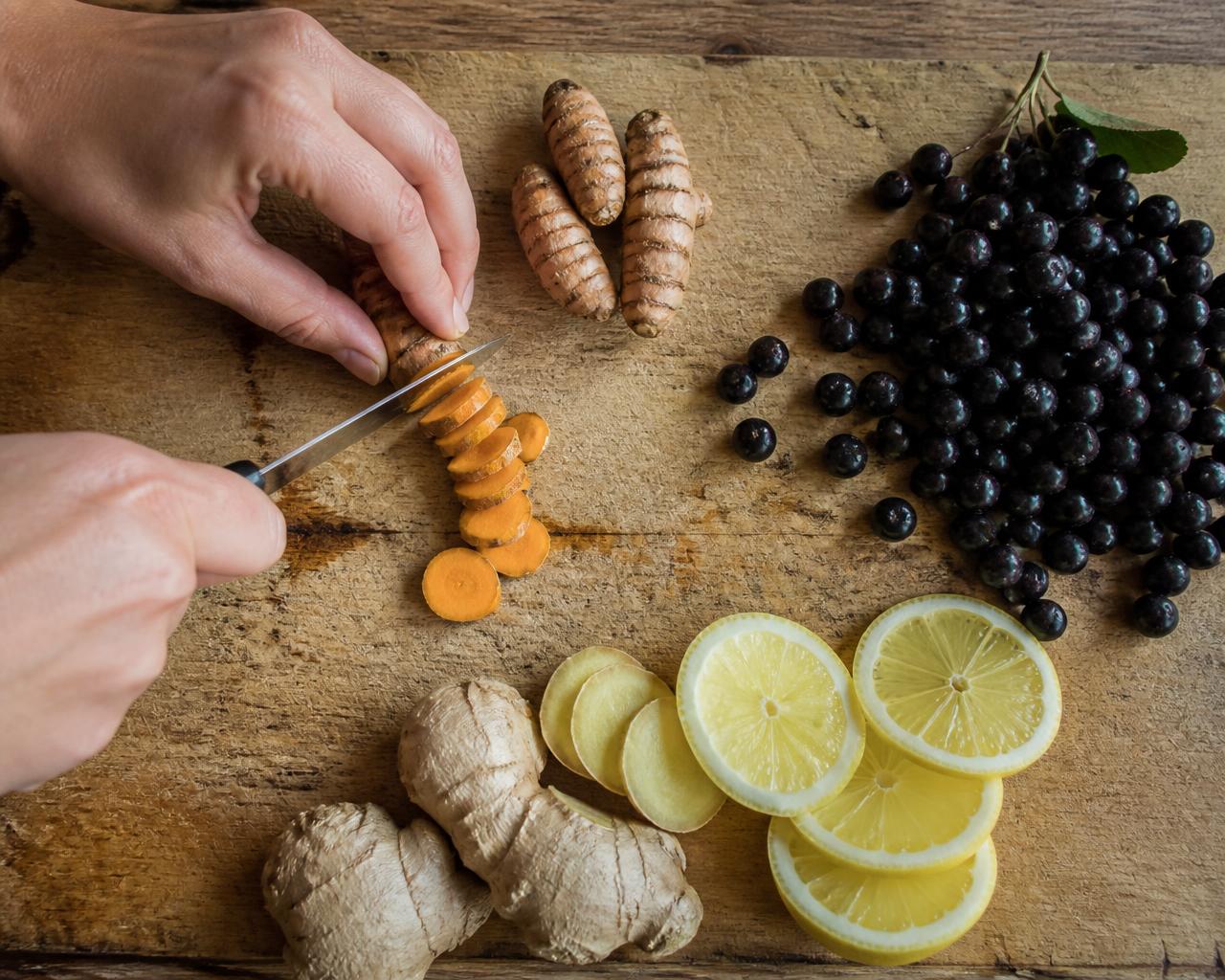 Hands slicing fresh turmeric, ginger and lemon beside dark Aronia berries